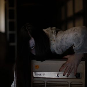 Cosplayerin verkleidet als das Mädchen aus dem Film "The Ring", blickt in die Kamera und klettert über einen alten Fernseher. Der Hintergrund ist sehr dunkel, das Mädchen in weißen, dreckigen Klamotten und mit weißem Make-Up im Gesicht, sticht heraus. Es handelt sich um ein Cosplayshooting.
