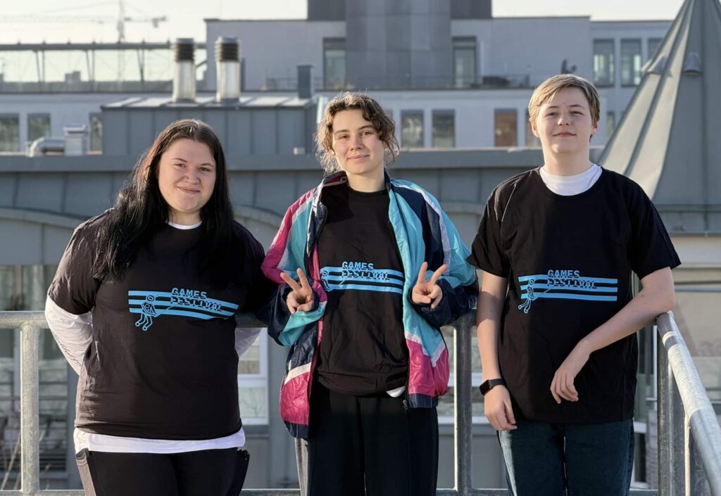 Drei junge Erwachsene stehen auf einem Balkon vor einem städtischen Hintergrund. Alle tragen schwarze T-Shirts mit einem blauen Design und dem Text "GamesFestival". Die Person in der Mitte zeigt ein Victory-Zeichen. Im Hintergrund sind Gebäude und ein Baukran zu sehen.